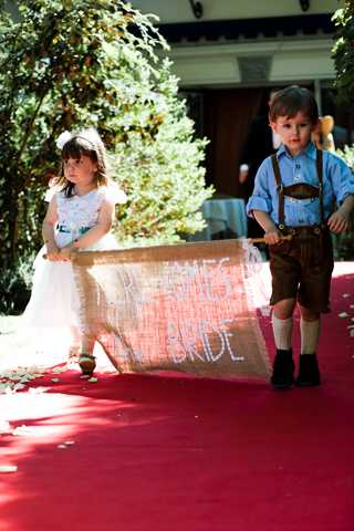 Two young children walk down a red carpet aisle during an outdoor wedding ceremony, carrying a large burlap banner with hand-lettered text reading 'BRIDE' and what appears to be a name above it. The girl on the left wears a white dress with a teal or blue sash and sandals, while the boy on the right wears a light blue shirt, leather-style brown suspender shorts (lederhosen style), knee-high white socks, and black shoes. The aisle is scattered with rose petals, and green hedges and a building facade are visible in the background. The image is a vertical portrait shot taken from a low angle, capturing the children walking toward the camera.
