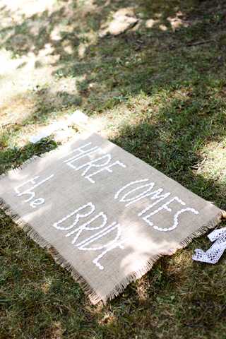 Detail shot of a rustic burlap sign reading 'Here Comes the Bride' in white hand-lettered text, laid flat on grass in dappled sunlight. The sign has frayed edges consistent with a rustic or boho styling theme. A small piece of white lace trim is visible at the lower right corner of the sign. The composition is a close-up flat lay taken from above.
