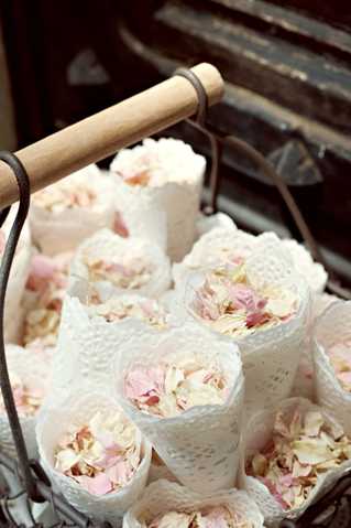 Close-up detail shot of a rustic wire basket with a wooden handle holding multiple cone-shaped pouches made from white lace doilies, each filled with dried flower petals in blush pink and cream tones. The cones appear to be confetti toss favors prepared for guests, likely for a ceremony exit. The styling is romantic and vintage-rustic, with the lace cones and soft petal colors creating a delicate, handcrafted aesthetic. The image has a slightly soft focus and warm, muted tones overall.