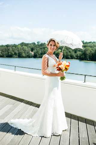 A bridal portrait taken outdoors on a wooden deck terrace, with a lake and tree line visible in the background. The bride stands alone holding a white lace parasol in one hand and a bouquet of orange, red, and yellow flowers — likely tulips, lilies, and ranunculus — in the other. She wears a fitted, floor-length white lace gown with a short train, and her hair is styled up with a small decorative accessory. The composition is a full-length portrait shot in natural daylight.