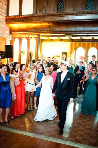 The bride and groom are making their entrance onto the dance floor at an indoor reception, surrounded by cheering guests on both sides. The groom wears a black tuxedo and a white wide-brim fedora-style hat, while the bride wears a white V-neck wedding gown. Guests are dressed in a colorful mix of attire including cobalt blue, coral, teal, and emerald green dresses. The venue features dark wood paneling, arched windows, and warm overhead lighting, suggesting a historic ballroom or chateau-style reception hall. The shot is taken from a wide angle at floor level, capturing the full crowd reaction as the couple walks in together.