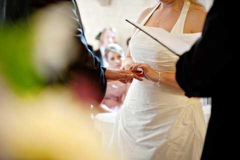 A close-up shot capturing the ring exchange moment during an indoor wedding ceremony. The bride, wearing a white halter-neck draped gown with a delicate necklace and pearl bracelet, places a ring on the groom's finger while an officiant holds a black folder nearby. A small group of seated guests is softly visible in the background, with one woman wearing a teal fascinator. The foreground features a blurred yellow floral arrangement, adding depth to the composition.