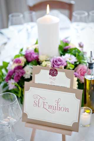 Close-up detail shot of a wedding reception table setting featuring a small wooden easel displaying a table name card reading 'St Emilion' in burgundy script on a cream and kraft paper card, with a matching menu card stacked behind it secured with a burgundy wax seal. The centerpiece in the background includes a large white pillar candle surrounded by a low floral arrangement of mauve roses, pink roses, cream blooms, and purple flowers with greenery. A wine glass and olive oil bottle are partially visible at the edges of the frame, suggesting a French-inspired reception styling with a classic, romantic decor palette of cream, burgundy, and shades of pink and purple.