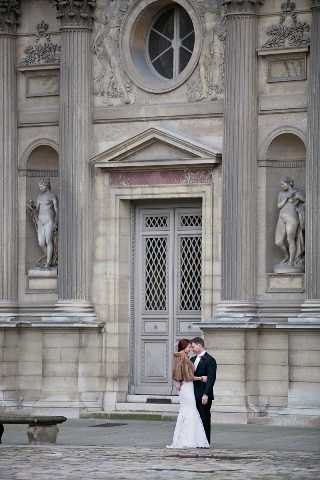 A bride and groom share an intimate moment standing in front of a grand classical French building, likely the Louvre in Paris, with ornate stone facades, pilasters, carved figural sculptures in niches, and a large arched doorway with diamond-patterned ironwork. The bride wears a fitted white gown with a fur stole or wrap in a warm caramel-brown tone, and the groom is dressed in a dark suit with a light tie. The couple is leaning into each other with their foreheads close, creating a romantic pose. This is a wide portrait shot that emphasizes the architectural grandeur of the backdrop against the small scale of the couple. Potential venue feature image.