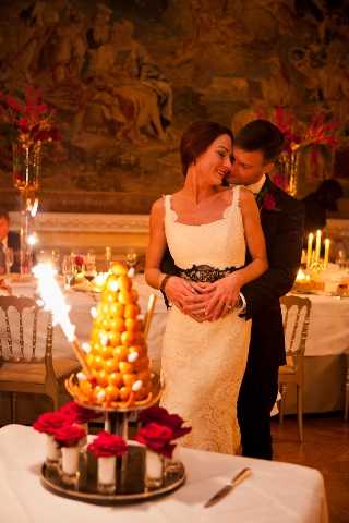 The bride and groom stand together behind a traditional French croquembouche displayed on a mirrored tray, decorated with red roses and lit with sparkler fountains. The scene takes place indoors in an ornate ballroom or chateau reception room featuring large tapestries on the walls, gold chiavari chairs, and candlelit tables with deep red floral centerpieces visible in the background. The bride wears a fitted ivory lace gown with a black lace sash at the waist, while the groom is dressed in a dark suit with a burgundy boutonniere; both are smiling and leaning into each other. This is a medium portrait shot with warm candlelight tones, consistent with a classic, formal French wedding reception aesthetic.