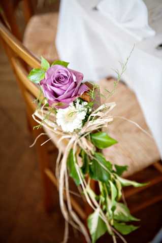 Close-up detail shot of a wooden ceremony chair decorated with a floral arrangement featuring a single lavender rose, a small white bloom, green foliage, and trailing natural raffia ribbon. The arrangement is tied to the back of a natural wood cross-back chair, with greenery cascading downward. A white fabric element, likely a chair sash or dress, is softly blurred in the background. The styling is rustic and understated, with a muted purple and ivory floral palette paired with natural materials.