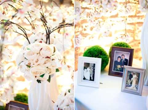 A side-by-side detail shot of wedding reception decor. On the left, a white vase holds a large bouquet of cream and ivory peonies and roses, set against a backdrop of faux cherry blossom branches with warm fairy lights creating a soft, glowing background. On the right, a memory table displays two framed photographs of previous generations' weddings — one black-and-white and one color — alongside a round green moss ball, all arranged on a white table surface with the same illuminated blossom branch backdrop behind them. The overall decor palette is ivory, cream, and white with warm ambient lighting, and the styling is classic and romantic. Both panels are close-up detail shots.