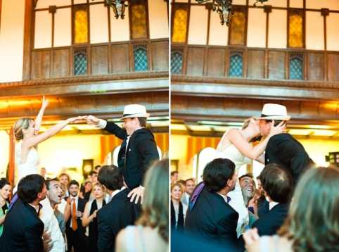 A side-by-side diptych showing a lively reception dance floor moment inside a grand indoor venue with Tudor-style architecture, warm amber lighting, and stained glass windows on an upper balcony level. In both frames, the bride — wearing a white strapless dress with her hair up — and the groom — in a black suit and white panama-style hat — are being lifted by a crowd of guests and appear to be dancing energetically together; in the left frame the bride raises her arm, and in the right frame the couple kisses while elevated. Approximately 10–15 guests are visible surrounding the couple, several in dark suits with one guest wearing an orange tie. The overall styling is classic with a festive, high-energy atmosphere, captured as a medium-wide shot.