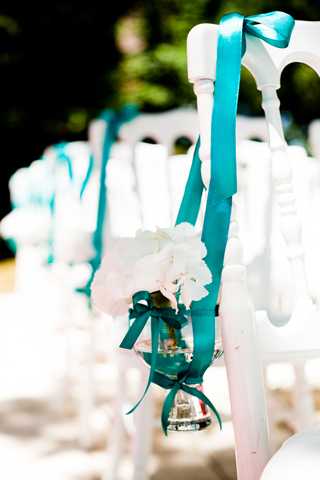 Close-up detail shot of outdoor wedding ceremony aisle chair decorations. White folding chairs are lined up along the aisle, each decorated with a wide teal satin ribbon tied in a bow at the top and a small glass votive holder with white florals — appearing to be white orchids — secured with a smaller teal ribbon bow at the base. The decor palette is teal and white, creating a crisp, clean color contrast. The background is softly blurred with additional decorated chairs visible receding into the distance.