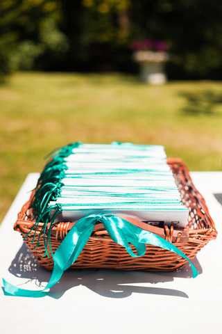 Close-up detail shot of a wicker basket holding a stack of wedding ceremony programs, each tied with a teal satin ribbon. The programs feature light blue or mint covers, and the teal ribbon accents create a cohesive color scheme. The basket is placed on a white surface, set outdoors in a garden setting with green foliage visible in the background. The overall decor palette suggests a teal and mint color theme for the wedding.