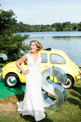 A bride stands outdoors laughing, posed in front of a bright yellow vintage Fiat 500 car parked near a lakeside setting. She wears a fitted white V-neck gown with a short train and has her hair styled in an updo. A large decorative metal letter 'S' stands beside her on the grass, used as a personalized decor prop. The shot is a full-length portrait with a casual, joyful feel against a backdrop of trees and open water.