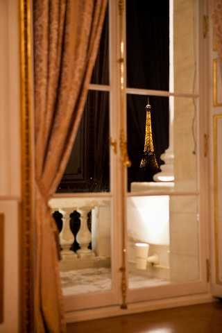 A detail shot taken from inside an ornate Parisian venue at night, looking through open French doors with gold-trimmed white molding toward the illuminated Eiffel Tower in the background. The interior features blush-toned heavy drape curtains and decorative wall paneling in the foreground, while a stone balustrade balcony is visible just outside the doors. No people are present in the frame — this is an atmospheric venue detail or establishing shot. Potential venue feature image.