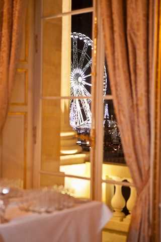 A detail shot taken from inside an indoor reception venue at night, looking through tall French doors framed by dusty rose/blush draped curtains toward an outdoor terrace and city view beyond. Through the glass panels, a large illuminated Ferris wheel and what appears to be the Eiffel Tower are visible against the night sky, suggesting a Paris city-center location. In the foreground, a white linen-covered table holds glassware, likely part of a cocktail hour or reception bar setup. The interior walls are warm golden-toned, consistent with classic Haussmann-style Parisian architecture. Potential venue feature image.