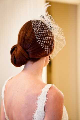 Close-up portrait shot taken from behind showing a bride's hair and accessory styling during a getting-ready moment indoors. The bride has deep auburn hair styled into a low, sleek chignon and is wearing a white birdcage veil with a small feathered fascinator clip at the crown. She wears a low-back white lace wedding dress with delicate frayed strap edges and a small drop earring is visible. The overall bridal styling aesthetic is vintage-inspired, referencing 1920s-1940s glamour through the birdcage veil choice.