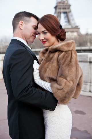 A couple portrait taken outdoors in Paris, with the Eiffel Tower visible in the background. The groom wears a dark navy or black suit with a white pocket square, while the bride wears a fitted white lace gown layered with a tan/camel-toned fur stole wrap. The bride has auburn hair styled in an updo with a decorative hair accessory and wears bold red lipstick. The two stand close together, foreheads nearly touching, in an intimate pose. The shot is a medium close-up portrait with a classic, vintage-inspired styling aesthetic.