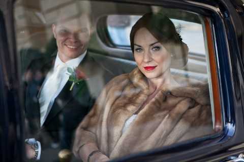 A close-up portrait of a bride and groom photographed through the window of a classic dark-colored wedding car. The bride is in the foreground wearing a tan fur stole over her wedding attire, with her dark hair styled in an updo accented by a small fascinator or hair piece, and she is wearing bold red lipstick. The groom is visible behind her, smiling, dressed in a dark suit with a white boutonniere featuring a red floral accent. The image has a vintage or 1940s-inspired styling aesthetic, reinforced by the retro vehicle, the fur wrap, and the bride's classic Hollywood makeup. The window glass creates a slight reflection overlay on the composition.
