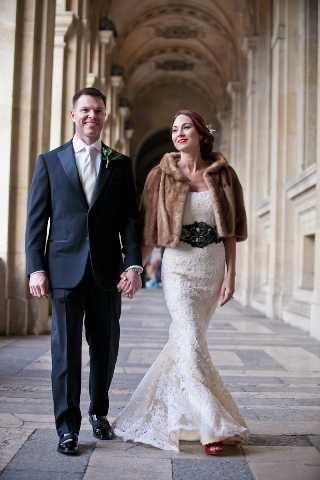 A couple portrait taken in a grand covered arcade or colonnade with ornate carved stone arches and vaulted ceiling visible in the background, likely in Paris. The bride wears a fitted lace mermaid-style gown with a black embellished belt and a brown fur stole draped over her shoulders, paired with red heels, while the groom wears a dark navy suit with a light tie and a green boutonniere. The two are walking hand in hand toward the camera, smiling. The composition is a mid-length portrait shot with the arched colonnade creating strong architectural perspective lines behind them. Potential venue feature image.
