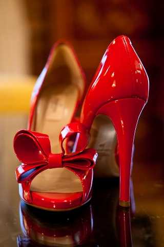 Close-up detail shot of a pair of red patent leather peep-toe platform heels with a bow embellishment at the toe, positioned on a reflective dark surface. The shoes feature a stiletto heel and glossy finish, likely bridal accessories. The warm, blurred background suggests an indoor setting. The image is sharply focused on the shoes, emphasizing them as a deliberate bridal styling choice.