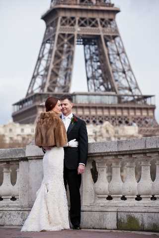 A couple poses for a wedding portrait on the Trocadéro esplanade in Paris, with the Eiffel Tower prominently visible in the background. The bride wears a fitted ivory lace mermaid-style gown with a brown fur stole draped over her shoulders, and white gloves, while the groom is dressed in a classic black tuxedo with a white boutonniere. The two face each other closely, standing against a stone balustrade railing. The shot is a medium-distance portrait taken at ground level, capturing both the couple and the iconic landmark in full.