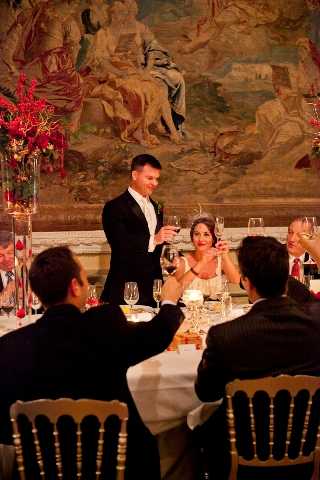 A toast is being made during a wedding reception dinner inside a grand ballroom or chateau dining room, with a large ornate tapestry depicting classical figures covering the wall behind the couple. The groom, dressed in a black tuxedo with a white boutonniere, stands and raises a wine glass while the bride, in a strapless white gown with dark hair, remains seated and raises her glass to meet his. Several guests are visible seated at the round dining table in the foreground, also raising glasses. The table is set with white linens, crystal wine glasses, candles, and tall floral centerpieces featuring deep red flowers and red berry branches. The room is warmly lit by candlelight and ambient lighting, creating a rich, golden tone throughout the image. The composition is a medium shot taken from slightly behind seated guests, framing the couple against the tapestry backdrop.