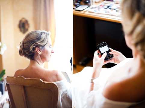 A diptych of two getting-ready images featuring the bride in an indoor setting, likely a hotel room or dressing room. In the left frame, the bride is seated in an ornate wooden chair with her back to the camera, wearing a white off-the-shoulder robe, her blonde hair styled in an upswept chignon. In the right frame, the bride is seen from behind holding a BlackBerry smartphone, with a vanity table covered in makeup products visible in the background. Both images have a candid, documentary feel with soft natural window light.