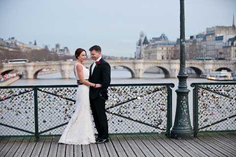 A bride and groom share a close, smiling moment on the Pont des Arts in Paris, with the Seine River, a stone bridge, and classic Parisian architecture visible in the background. The bride wears a fitted white lace gown with a sleeveless neckline, while the groom is dressed in a black tuxedo with a red boutonniere. The bridge railing is densely covered with padlocks, a well-known feature of this location. This is a medium wide portrait shot taken at ground level on the wooden deck of the bridge.