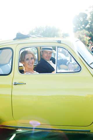 A bride and groom are seated inside a vintage yellow-green compact car, likely a classic Fiat 500 or similar retro vehicle, photographed from outside the passenger window. The bride, wearing a white dress with her hair up, smiles toward the camera, while the groom, dressed in a dark suit and a white flat cap, sits in the driver's seat. The shot is a close-up portrait framed by the car window, with strong backlighting creating a bright, sun-flared outdoor atmosphere. The vintage car serves as a styled transportation or decor element, adding a retro, playful touch to the wedding aesthetic.