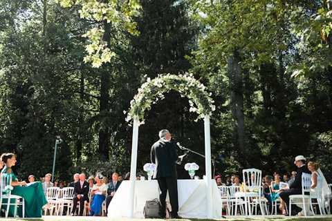 An outdoor wedding ceremony taking place in a wooded garden setting, with guests seated in white Chiavari chairs arranged in rows on either side of a central aisle. A white arch structure decorated with white and green floral arrangements — appearing to include white blooms and trailing greenery — frames the altar area at the center. An officiant stands at a microphone facing the seated guests, with a white cloth-covered table nearby. Approximately 30–40 guests are visible, dressed in a mix of formal attire including teal and blue dresses. The ceremony has a classic, garden-party aesthetic with an all-white decor palette. Wide shot taken from behind the officiant, capturing the full ceremony layout and surrounding tall trees.