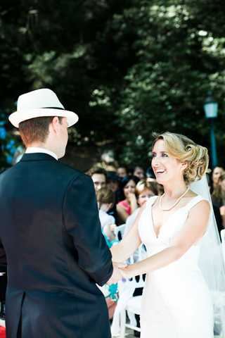 An outdoor wedding ceremony showing the bride and groom facing each other and holding hands at the altar. The groom wears a dark navy suit with a white fedora-style hat, while the bride wears a fitted white gown with a V-neckline, a pearl necklace, and a veil, her blonde hair styled in an updo. The bride is laughing and smiling broadly toward the groom. Rows of white chairs filled with approximately 20 or more guests are visible in the background, set beneath leafy trees. The overall styling is classic with a relaxed, summery feel. This is a medium portrait shot taken from slightly behind the groom, focusing on the bride's expression.