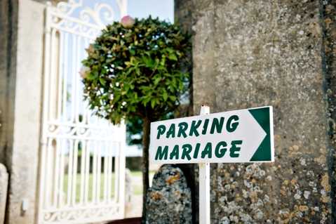A close-up detail shot of a green and white directional sign reading 'PARKING MARIAGE' with a right-pointing arrow, mounted on a white post beside a stone wall. In the soft-focus background, a white ornate wrought-iron gate is visible alongside a neatly trimmed topiary ball tree. The sign uses dark green lettering on a white background with a green border, consistent with classic French roadside signage styling.