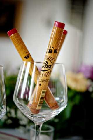 Close-up detail shot of two cigars with red tips and branded wooden-look paper tubes, placed upright inside a clear crystal wine glass, likely set out as a guest favor or welcome gift at a reception table. The branding on the tubes reads 'Puros' and includes additional Spanish text suggesting authentic cigars. Soft floral arrangements in the background are slightly out of focus, with white and green tones visible. The overall styling suggests an indoor reception setting with a relaxed, festive atmosphere.