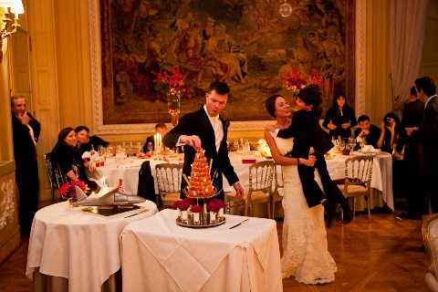 A couple cuts into a traditional French croquembouche — a tower of cream puffs — at their wedding reception, with the groom in a black tuxedo leaning in while the bride in a fitted ivory gown embraces him playfully. The setting is an ornate ballroom with warm golden lighting, gilt wall paneling, and a large classical tapestry or oil painting mounted on the wall behind them. Guests seated at white linen-covered tables in the background are dressed in formal black attire, with red floral centerpieces providing accent color throughout the room. The composition is a medium wide shot capturing the cake-cutting moment within the full reception context.