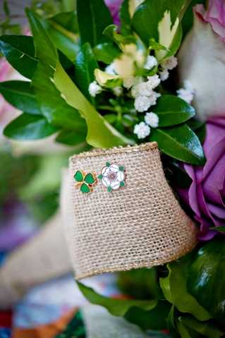 Close-up detail shot of a bridal bouquet wrapped in a natural burlap ribbon, with two decorative enamel pins attached to the wrap — one shaped like a green and gold four-leaf clover and one depicting a white flower with green and pink accents. The bouquet includes purple/mauve roses, small white cluster blooms, and lush green foliage. The burlap wrap gives the styling a rustic, countryside feel. The background is softly blurred with hints of additional floral colors.