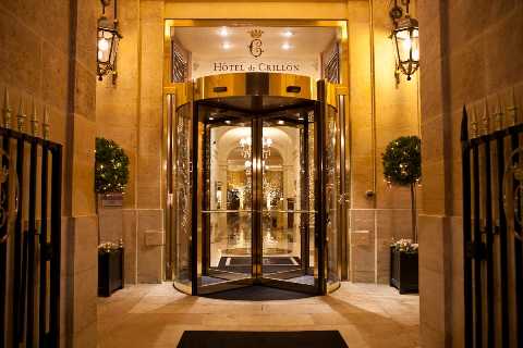 This image shows the entrance of the Hôtel de Crillon in Paris, featuring a brass-framed revolving door set within a carved stone facade, lit by ornate wall lanterns casting warm golden light. No wedding party or guests are visible; the shot captures the hotel's exterior entrance at night with topiary boxwood plants in dark planters flanking the doorway and a chandelier visible through the glass interior. The composition is a straight-on wide shot of the entryway. Potential venue feature image.