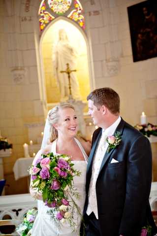 A bride and groom share a moment together inside a Catholic church following their ceremony, positioned in front of the main altar. The bride wears a strapless white gown with a veil and holds a cascading bouquet of purple and mauve roses with green foliage. The groom is dressed in a dark suit with a cream waistcoat and a buttonhole featuring purple flowers and greenery. In the background, a white marble statue of a religious figure is illuminated within an arched niche, flanked by stained glass windows and candles on the altar. The portrait-style shot is taken at a slight angle with shallow depth of field, keeping the couple in focus while the altar setting is softly blurred.