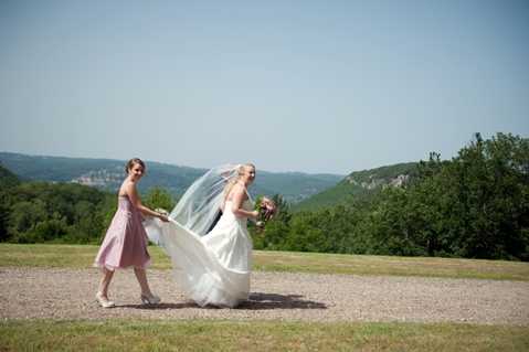 A bride and one bridesmaid are photographed outdoors on a gravel surface with an open lawn area behind them. The bridesmaid, wearing a knee-length dusty pink dress, is holding the train of the bride's white ballgown as it billows in the wind, while the bride holds a small bouquet and wears a long veil. The setting appears to be an elevated outdoor area with forested hillsides visible in the background. The shot is a full-length wide portrait captured in natural daylight.