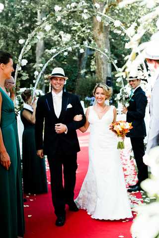 The bride and groom walk back down the aisle together following an outdoor ceremony, arm in arm and smiling at guests. The groom wears a black suit with a white dress shirt, bow tie, and a white fedora-style hat, while the bride wears a fitted V-neck lace gown and carries a bouquet of orange, red, and yellow flowers. The aisle is lined with a red carpet scattered with white rose petals, and a circular floral arch decorated with white flowers frames the couple in the background. Bridesmaids in floor-length teal dresses and guests in semi-formal attire line both sides of the aisle, with at least one male guest also wearing a white hat, suggesting a coordinated vintage or Gatsby-inspired styling theme. This is a medium portrait shot taken outdoors under tree cover.