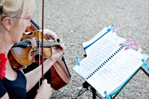 A close-up shot of a female violinist performing outdoors at a wedding, likely during a ceremony or cocktail hour. She is wearing a black top and has a red flower pinned to her outfit, and is reading from sheet music on a stand secured with colorful blue and pink clips. The image is taken from slightly above and behind, focusing on the violin, bow, and music stand in the foreground.
