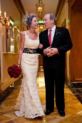 A bride and an older man, likely her father, walk together arm-in-arm along an interior corridor with warm gold tones, ornate wall sconces, and a polished marble floor, suggesting a classic hotel or formal venue. The bride wears a fitted ivory lace gown with a black sash at the waist, paired with a white birdcage veil and a black fascinator, and carries a compact bouquet of deep red roses. The man wears a black suit with a red tie and a dark red boutonniere. The portrait-style shot is taken at mid-length in warm artificial lighting.