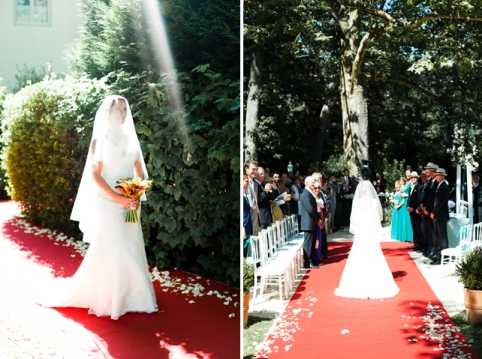 A diptych of two outdoor ceremony images showing the bride walking down the aisle on a red carpet runner scattered with white flower petals. In the left frame, a close-up portrait shows the bride in a fitted white gown with a long cathedral veil, holding a bouquet of warm-toned orange and yellow flowers, with a shaft of sunlight visible overhead. In the right frame, a wider shot captures the bride being escorted down the aisle past rows of white chairs filled with approximately 20-30 seated guests dressed in formal attire, set within a garden or chateau grounds shaded by large trees. The ceremony setup follows a classic, formal style with the red aisle runner and white seating arrangement.