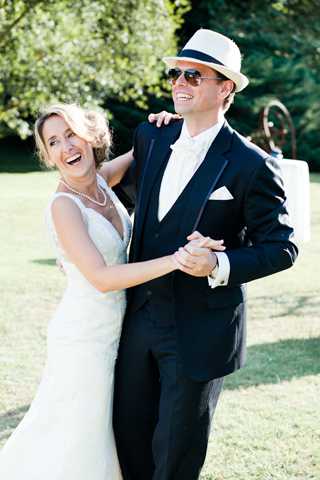 A bride and groom share a lighthearted, laughing moment together outdoors on a lawn, appearing to dance or pose playfully. The bride wears a fitted ivory lace spaghetti-strap gown and a pearl necklace, with her blonde hair pinned up. The groom is dressed in a dark navy suit with a white tie and pocket square, wearing aviator sunglasses and a white fedora with a navy band. The portrait-style shot is taken in natural daylight with a soft green garden background, capturing a candid, joyful interaction between the couple.