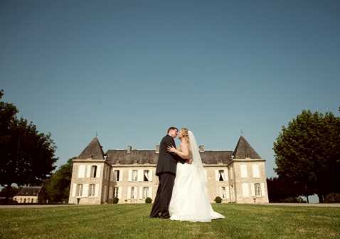 A bride and groom share a kiss or close embrace on a wide lawn directly in front of a French chateau, positioned at the center of the frame in a wide-angle portrait shot. The bride wears a white ball gown with a long cathedral-length veil, while the groom is dressed in a dark suit. The chateau behind them is a classic French country-style building with grey slate turret roofs, cream stone facade, and symmetrical windows across two stories. The composition places the couple small against the grand scale of the building and grounds, emphasizing the architectural setting. Potential venue feature image.