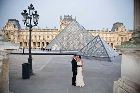 A bride and groom share a kiss during a couples portrait session in the Cour Napoléon at the Louvre in Paris, with the iconic glass pyramid prominently visible in the background along with the classical façade of the museum. The bride wears a white strapless floor-length gown and the groom is dressed in a black tuxedo. The wide shot captures the full architectural context, including a traditional Parisian street lamp in the foreground and a small crowd of visitors visible near the pyramid in the distance. The image has a classic, clean aesthetic with an early evening light casting soft, even tones across the plaza.