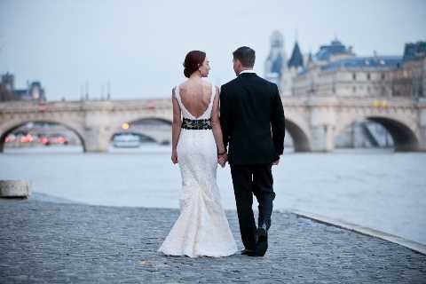 A couple portrait shot taken outdoors along the Seine river in Paris, with the Pont Neuf bridge and city buildings visible in the background. The bride wears a fitted, low-back lace gown with a black belt or sash detail at the waist, and the groom is dressed in a black tuxedo; they are holding hands and walking away from the camera. The composition is a medium-wide shot taken from behind, emphasizing the open back of the bride's dress and the Parisian cityscape. The overall styling is classic and formal, with muted, cool-toned ambient light suggesting dusk or overcast conditions.