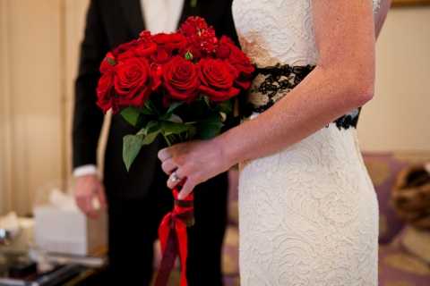 A close-up portrait shot focusing on the torso of a bride holding a tightly clustered bouquet of deep red roses tied with a red ribbon, set against a softly blurred indoor background. The bride is wearing a fitted white lace dress with a distinctive black lace sash or belt detail at the waist, adding a bold contrast to the white gown. Behind her, partially visible, is a groom in a dark suit with a white dress shirt. The decor palette is a striking red and black combination against white, suggesting a bold, high-contrast styling theme.