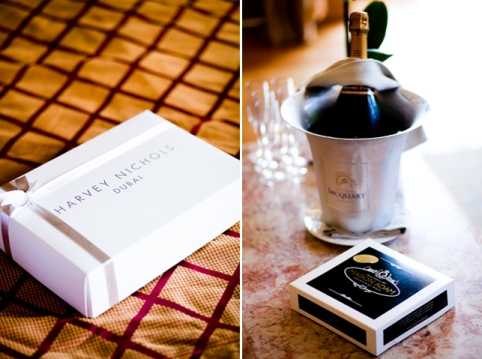 A close-up detail diptych showing two getting-ready vignettes. On the left, a white gift box branded 'Harvey Nichols Dubai' with a white ribbon sits on a patterned gold and burgundy fabric surface. On the right, a bottle of Jacquart champagne chills in a silver ice bucket alongside two champagne flutes and a branded matchbox, arranged on a marble surface. The images are detail shots with a shallow depth of field, suggesting pre-ceremony preparations in an indoor setting with a classic styling approach.
