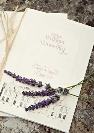A close-up flat lay of a wedding ceremony booklet printed in dusty rose typography on cream card stock, reading 'Wedding Ceremony, Chateau de Lognac, 25th June 2012,' with a hand-illustrated sketch of a French chateau printed at the bottom of the cover. Two sprigs of fresh purple lavender are laid across the booklet, and a second booklet tied with natural raffia ribbon is partially visible behind it. The shot is styled against a weathered stone surface, suggesting a Provençal or South of France aesthetic.