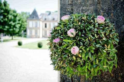 Close-up detail shot of a spherical floral arrangement mounted on a stone pillar or wall, featuring lush green foliage densely packed with soft pink garden roses or ranunculus blooms. The arrangement appears to be used as an entrance or ceremonial decoration. In the soft-focus background, a classic French chateau with grey slate roofing is visible, along with a formal gravel courtyard and manicured topiary balls. Potential venue feature image.