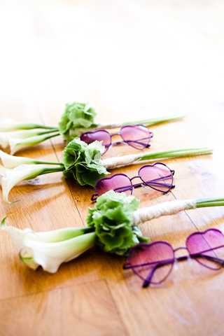 A detail flat lay shot arranged on a warm wood surface, featuring three white calla lily boutonnieres wrapped with twine, each paired with a heart-shaped purple-tinted sunglasses as a fun wedding accessory. The boutonnieres include small green blooms alongside the calla lilies. The image is shot with a shallow depth of field, with the foreground in sharp focus and the background softly blurred, giving a whimsical, playful styling aesthetic to what appears to be a bridal party or guest accessory arrangement.