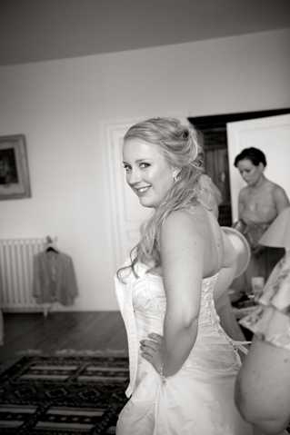 A black-and-white getting-ready scene shot indoors in what appears to be a room within a French chateau or manor house. The bride, a blonde woman with her hair in a loose updo, stands in the foreground wearing a strapless fitted wedding gown with lace or embroidered bodice detail, smiling over her shoulder toward the camera. In the background, a bridesmaid or helper in a strapless top appears to be assisting with the dress, while a suit jacket or robe hangs on a rack near the wall. The image has moderate contrast with soft mid-tones, and is composed as a candid portrait shot from a medium distance.